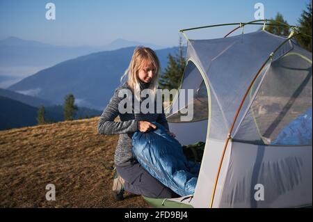 Young woman sitting down near her tent set up on grass lawn and laying out sleeping bag on the background of silhouettes of mountain hills. Autumn camping in the mountains. Stock Photo