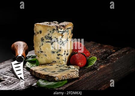 Semi-soft cheese with mold made from cows milk. The king of blue cheeses is Stilton. A piece of cheese and ripe strawberries on a dark wooden board an Stock Photo