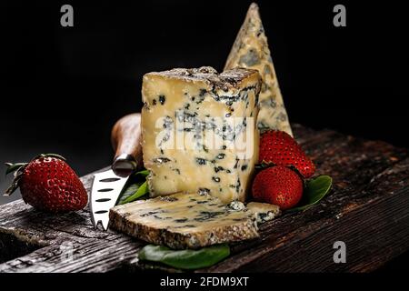 Semi-soft cheese with mold made from cows milk. The king of blue cheeses is Stilton. A piece of cheese and ripe strawberries on a dark wooden board an Stock Photo
