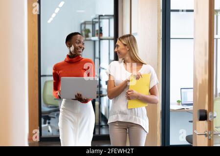 Happy diverse female business colleagues walking through corridor holding documents and talking Stock Photo