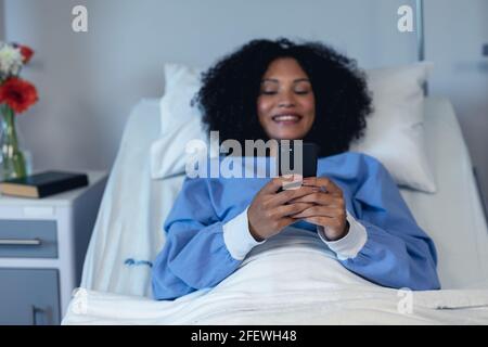 Smiling african american female patient lying in hospital bed using smartphone Stock Photo