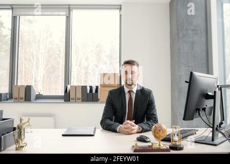 Portrait of serious handsome young executive in black suit and tie sitting with clasped hands at table equipped with computer Stock Photo