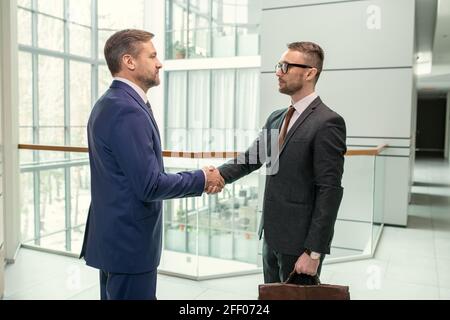 Side view of successful businessmen in suits standing in contemporary hall and shaking hands after signing contract Stock Photo