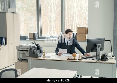 Concentrated young manager in formal suit sitting at desk and using computer while examining document in office Stock Photo