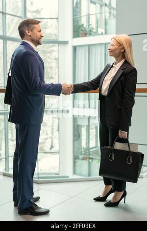 Smiling business partners in suits standing in corridor and shaking hands while congratulating each other with contract Stock Photo