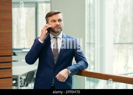 Serious thoughtful middle-aged businessman with stubble leaning on railing in office corridor and talking on phone Stock Photo