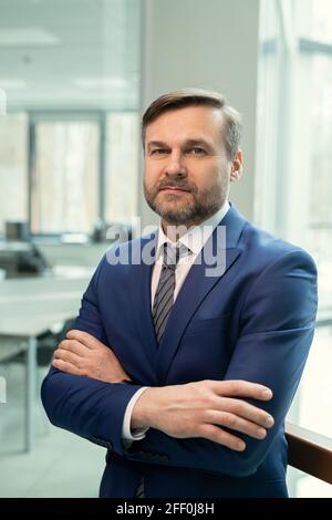 Portrait of content bearded manager in dark blue suit standing with crossed arms confidently against office room Stock Photo