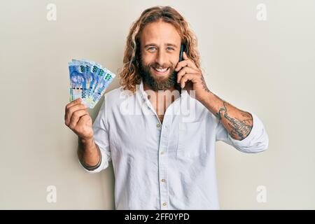 Handsome man with beard and long hair talking on the phone holding 100 brazilian reals smiling and laughing hard out loud because funny crazy joke. Stock Photo