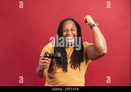 Excited African-American guy holds joystick isolated on red background, celebrating victory, overjoyed black guy won in video game on the console, screams yes and raising fist in triumph gesture Stock Photo