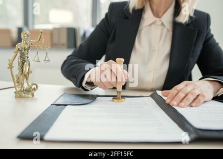 Close-up of unrecognizable female lawyer in black suit sitting at desk with themis statute and stamping document Stock Photo