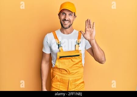 Young handsome man wearing handyman uniform over yellow background waiving saying hello happy and smiling, friendly welcome gesture Stock Photo