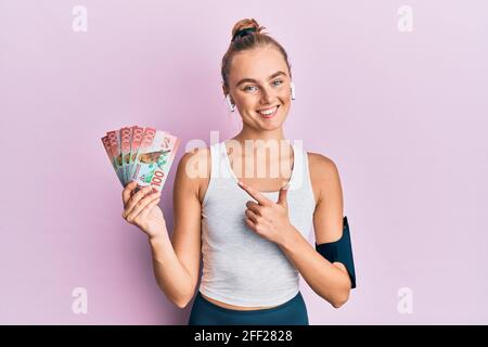 Beautiful blonde sport woman holding 100 new zealand dollars banknote smiling happy pointing with hand and finger Stock Photo