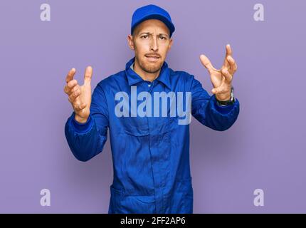 Bald man with beard wearing builder jumpsuit uniform shouting frustrated with rage, hands trying to strangle, yelling mad Stock Photo