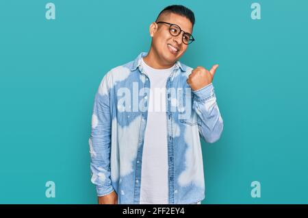 Young latin man wearing casual clothes and glasses smiling with happy face looking and pointing to the side with thumb up. Stock Photo