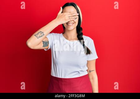 Young hispanic woman wearing professional waitress apron smiling and laughing with hand on face covering eyes for surprise. blind concept. Stock Photo