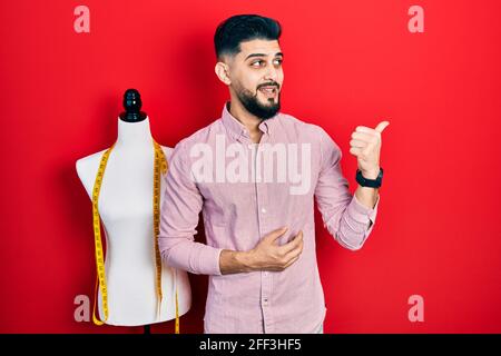 Handsome tailor man with beard standing by manikin pointing thumb up to the side smiling happy with open mouth Stock Photo