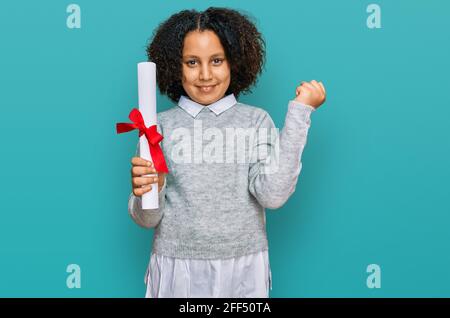 Young little girl with afro hair holding graduate degree diploma screaming proud, celebrating victory and success very excited with raised arms Stock Photo