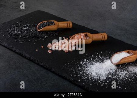 Different types of salt. White sea, pink himalayan and black volcanic salt. Three wooden spoons on black stone surface. Stock Photo
