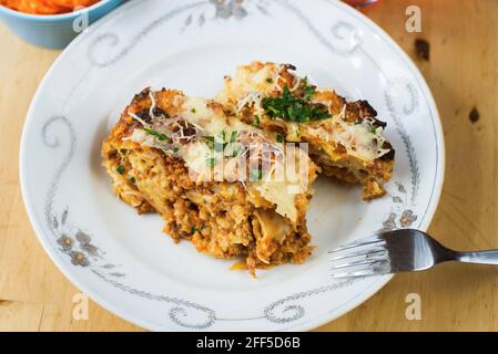 Baked pasta with minced meat gratinated with cheese sprinkled with fresh green parsley on white plate with fork on wooden table, rural meal. Stock Photo