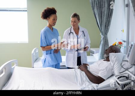 Two diverse female doctors and african american male patient in hospital room talking Stock Photo