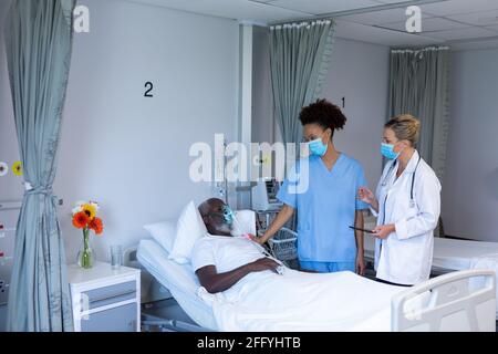 Two diverse female doctors wearing masks standing next to male patient in hospital room Stock Photo