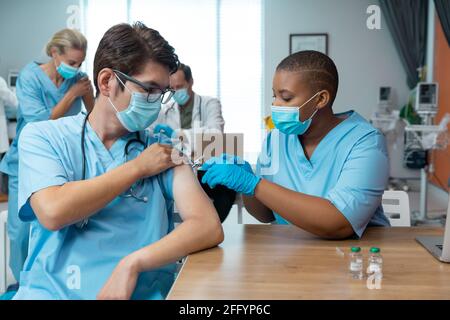 African american female doctor giving covid vaccination to male colleague, both in face masks Stock Photo
