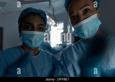 Diverse female doctors wearing face masks and surgical overalls looking at camera Stock Photo
