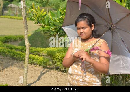 Teenage Indian Bengali girl holding an umbrella on her shoulder posing for photo in a garden, selective focusing Stock Photo