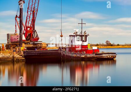 A small rustic vintage tow boat with chipped paint docked by a crane on Nanticoke river. A long exposure fine art image with silky smooth water and ru Stock Photo