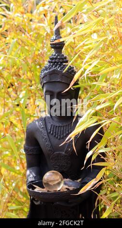 Standing black garden Buddha with a saucer in both hands. There is a glass ball in the saucer. Behind the Buddha yellow bamboo is growing Stock Photo