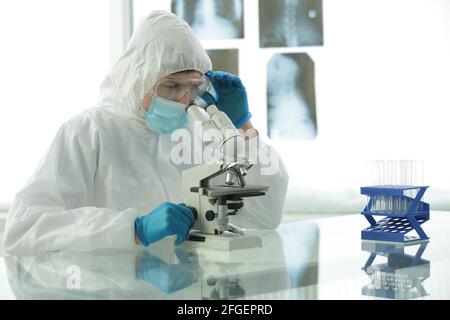 Doctor in a protective medical suit with gloves adjusts protective glasses near a microscope in a medical laboratory Stock Photo