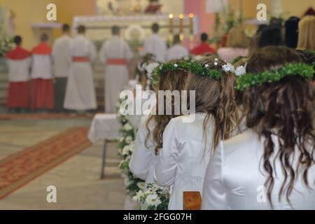Traditional celebration Fisrt Holy Communion in Catholic church in Poalnd, young girls in white dresses and wreath during holy mass. Stock Photo