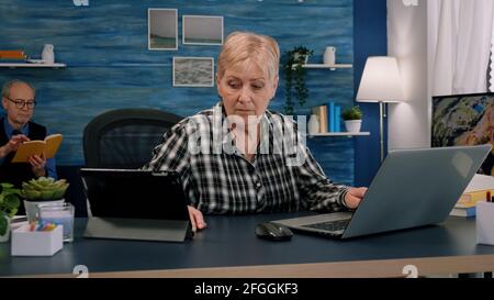 Old manager woman using laptop and tablet in same time working in home workspace for start-up company. Multitasking employee using modern technology network wireless overworking writing, searching Stock Photo