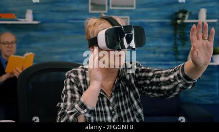 Retired concentrated senior woman using virtual reality goggles in living room gesturing sitting in home workplace. Senior woman experiencing vr headset while man sitting on sofa and reading a book Stock Photo