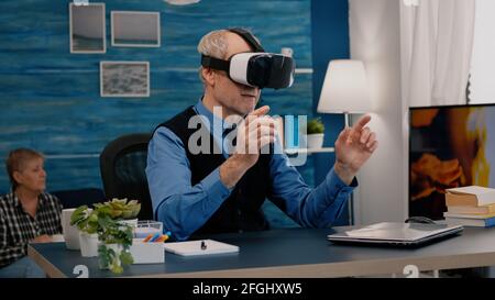 Retired man experiencing virtual reality using vr headset in living room working from home. Old remote employee searching, analysing financial reports while elderly wife watching tv in background Stock Photo