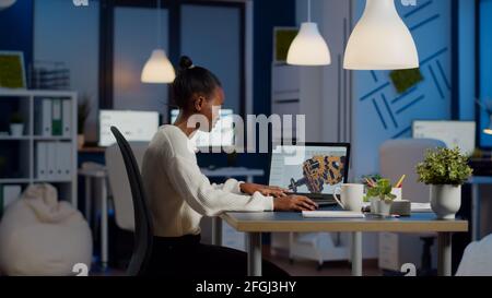 African engineer working late at night on a 3D model of industrial gears at laptop doing overtime in start up office. Freelancer studying prototype idea on pc showing cad software on device display Stock Photo