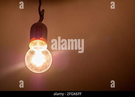 One single old plain bright incandescent lightbulb on in a darkened room, object detail, closeup, from below Bright yellow light source illuminates th Stock Photo