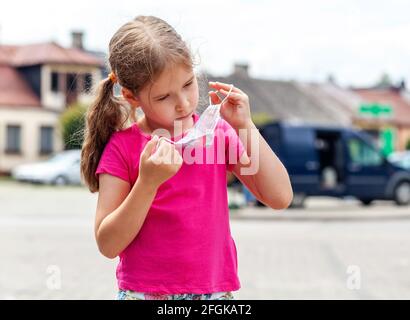 Young little school age girl, child putting on her protective medical face mask, outdoors portrait. Children and covid 19, corona virus protection lif Stock Photo