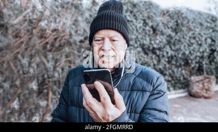 Smiling older man in cap and coat making a video call with his black cell phone and white helmets in the green garden Stock Photo