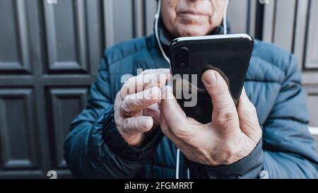 Hands of an elderly man holding his smart phone making a video call with his white headphones outdoors Stock Photo