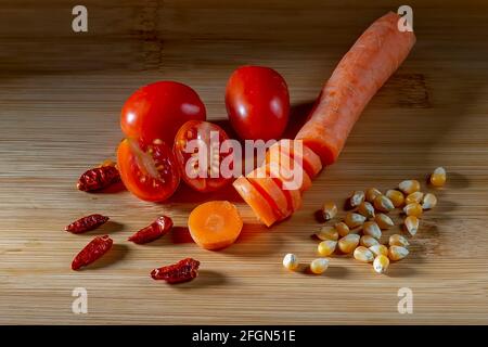 Partially sliced red cherry tomatoes and a carrot on a wooden cutting board, with red chillies and corn kernels Stock Photo