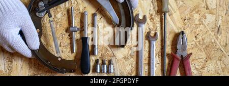 car drum brake disassembled in men's gloved hands and a set of various repair hand tools or auto mechanic's tools kit on a sheet of osb plywood backgr Stock Photo