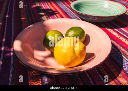 image of pink and pastel green soup plates, two lemons, and lime on a colorful table cloth shot in the sunlight Stock Photo