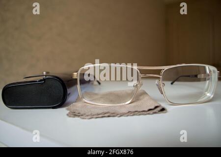 Glasses in gold metal frames lie on white wooden nightstand, next to glasses case and brown microfiber cloth against a blurred wall and door. Concept: Stock Photo