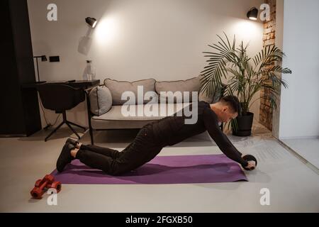 Workout at home. Sports and everyday life. Young man doing exercises on the press with a roller. He is dressed in a tracksuit, there are dumbbells and Stock Photo