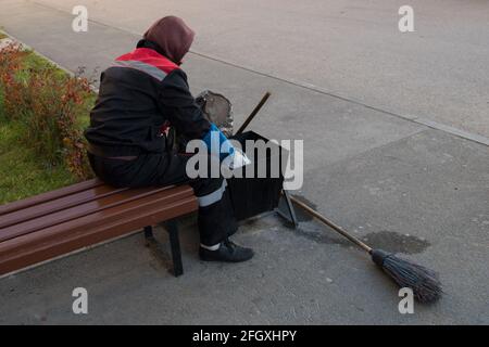 A man janitor in uniform sitting on wooden brown bench collects by hands in gloves garbage from black trash basket in a plastic bag, behind the basket Stock Photo