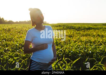 Pregnant happy young woman in the sunshine at sunset on nature holds her stomach in her hands and smiles Stock Photo
