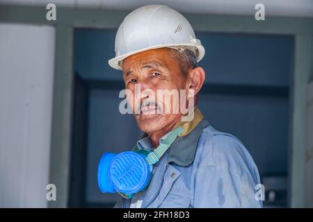 Gold ore mining and processing plant Altynalmas. Portrait of posing senior Asian worker in white hardhat with respirator mask. Almaty region. Stock Photo