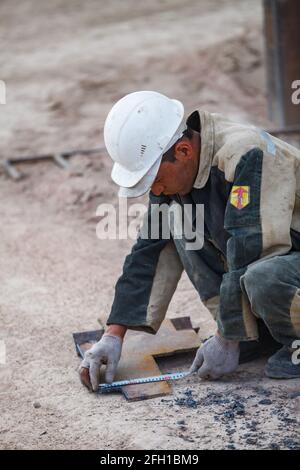 Mining and processing plant Altynalmas. Asian worker in white hardhat measuring metal part by measuring tape. Almaty region, Kazakhstan Stock Photo