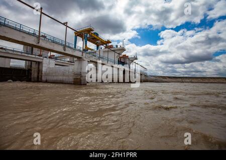 Hydraulic engineering. Shardara river dam near Kazaly city. Overhead crane on bridge.Blue sky with white clouds.Yellow river water.Kyzylorda region.KZ. Stock Photo